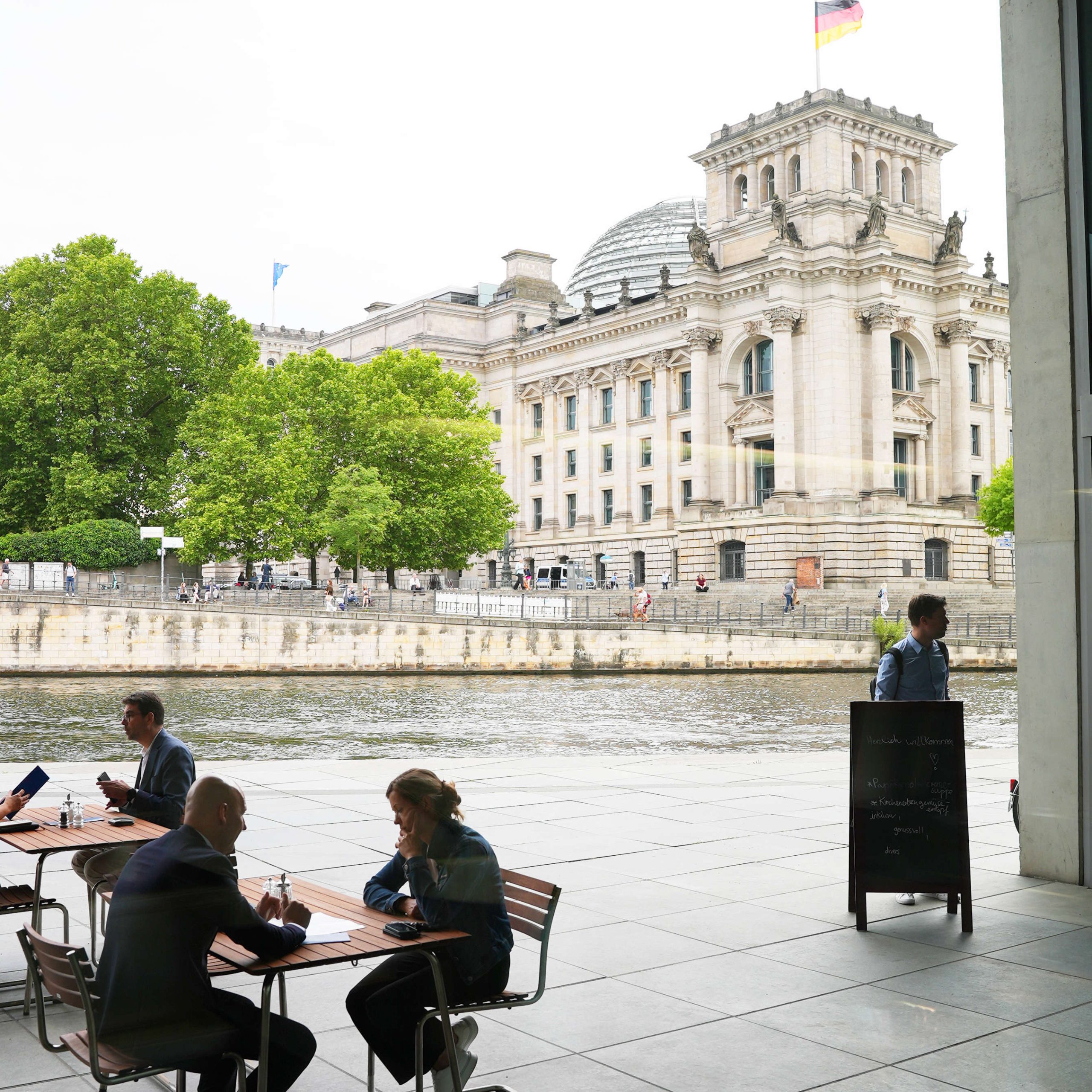 Gäste sitzen an Tischen des Bistro Marie. Im Hintergrund sehen wir die Spree und das Reichstagsgebäude.